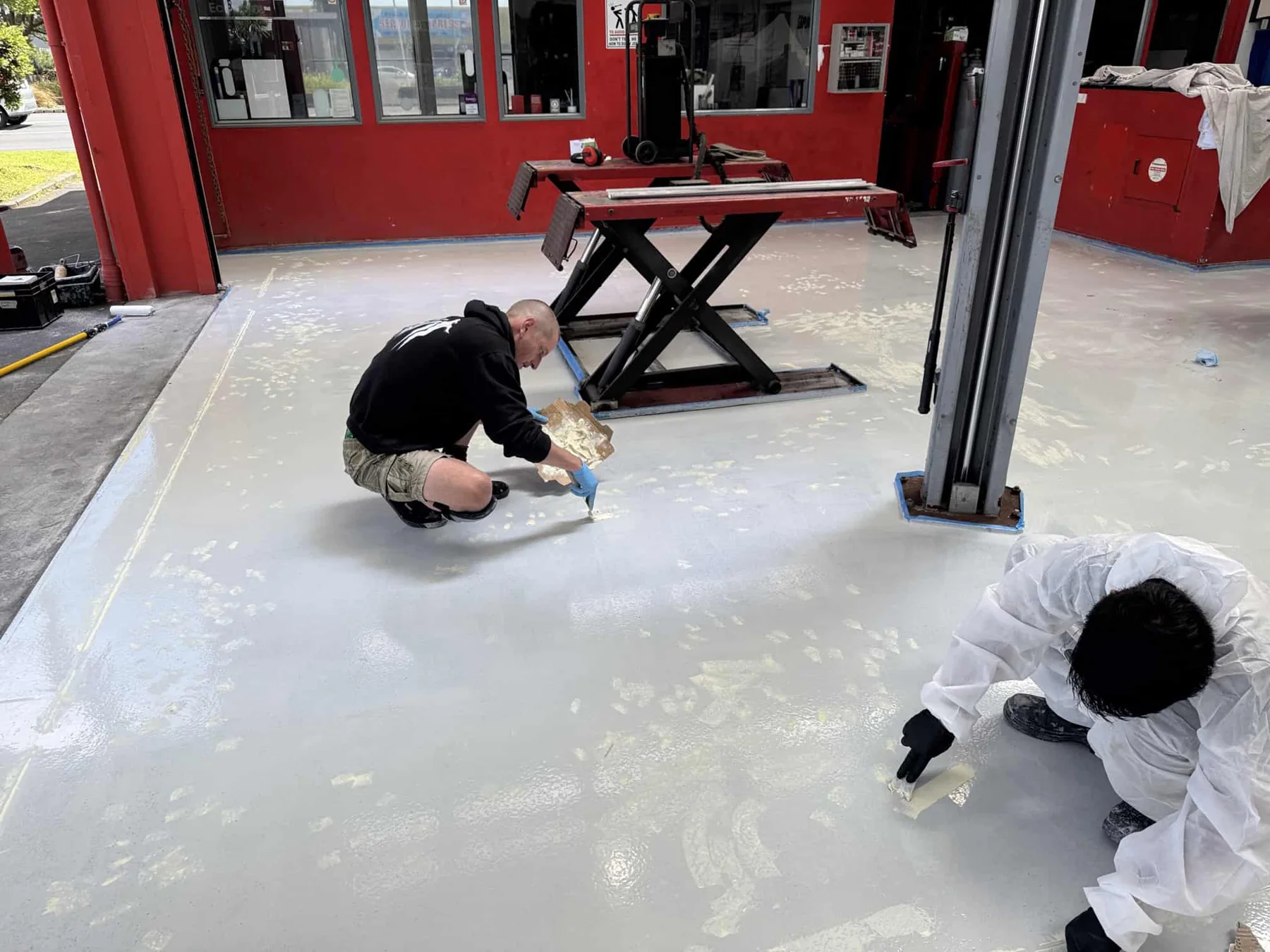 Flooring contractors applying a light-grey epoxy floor coating around workshop hoists